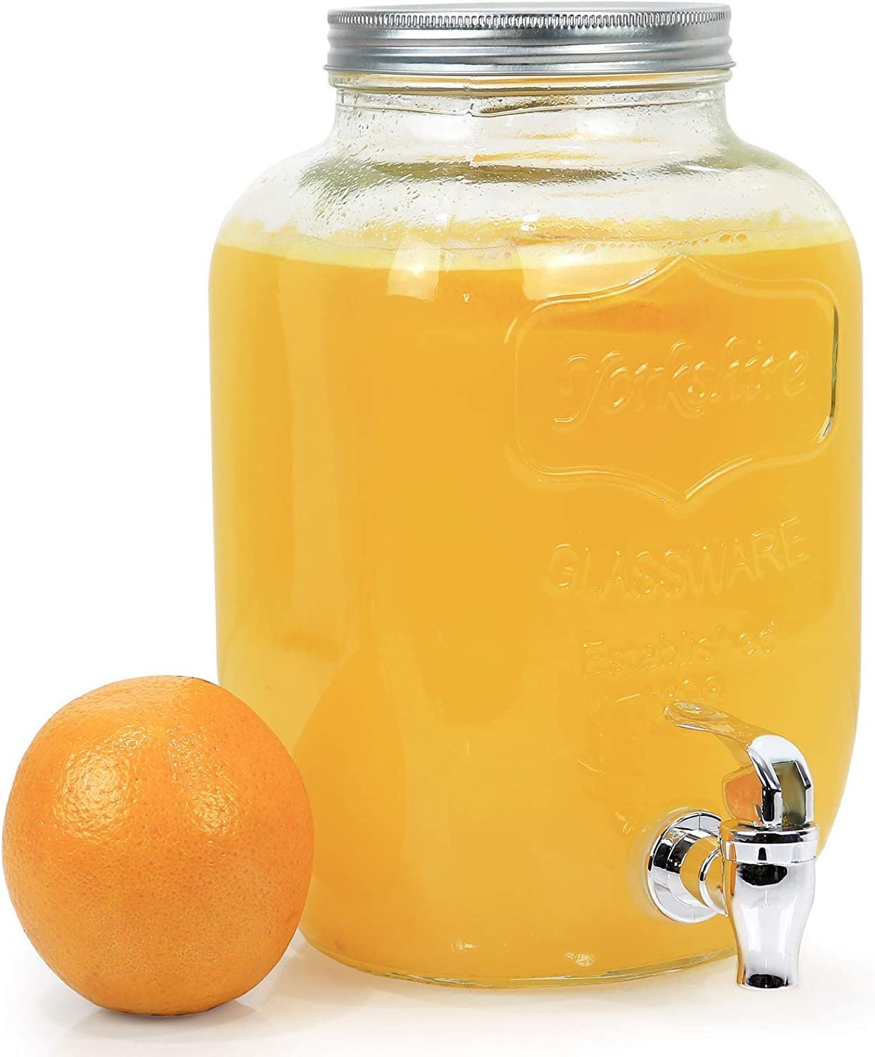 Glass drink dispenser filled with orange juice, with orange beside it